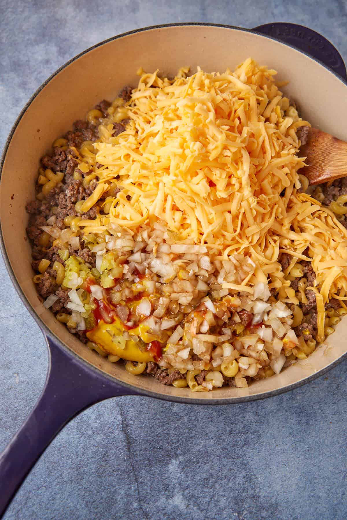 A skillet contains a mix of ground beef, macaroni, diced onions, shredded cheddar cheese, ketchup, and mustard—creating a deconstructed cheeseburger macaroni dish. A wooden spoon is placed inside the skillet. The ingredients are not yet mixed, showcasing their individual textures and colors.