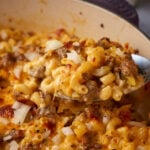 A close-up of a spoonful of cheesy casserole. The dish contains cooked elbow macaroni, ground meat, and diced onions, all mixed together and covered in melted cheese. The background shows more of the casserole in the dish.