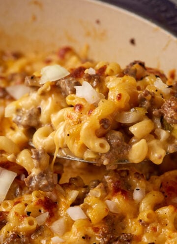 A close-up of a spoonful of cheesy casserole. The dish contains cooked elbow macaroni, ground meat, and diced onions, all mixed together and covered in melted cheese. The background shows more of the casserole in the dish.