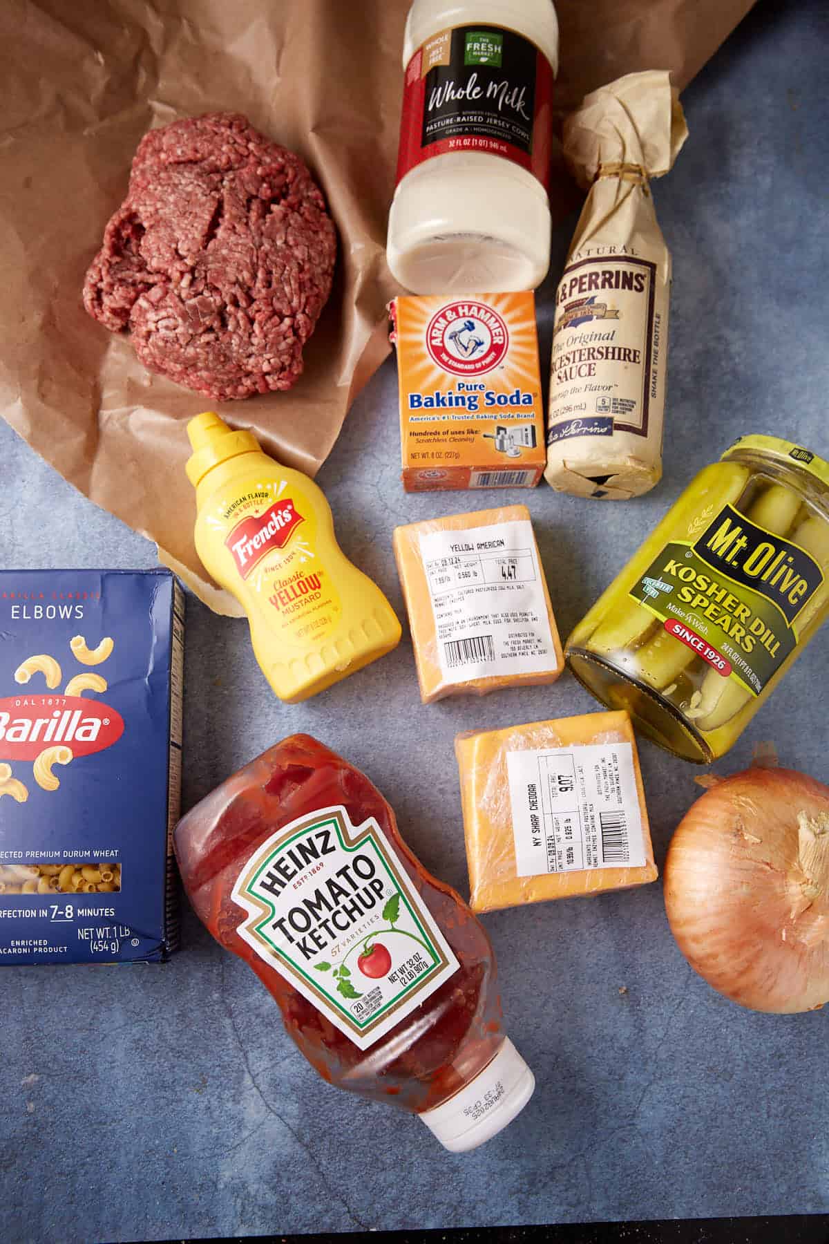 Various ingredients are laid out on a surface: ground beef, a bottle of whole milk, Worcestershire sauce, baking soda, pickle spears, a yellow onion, blocks of cheese, tomato ketchup, mustard, and a box of Barilla elbow pasta—all the makings for a delicious cheeseburger macaroni.
