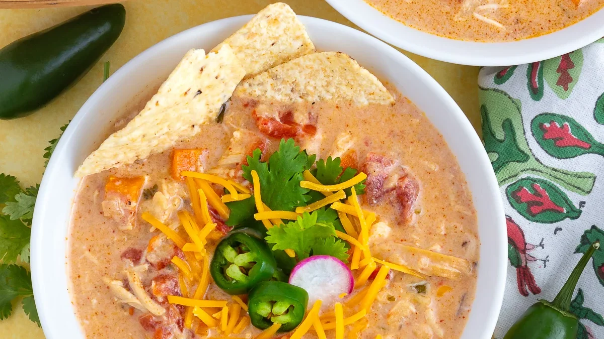 A bowl of creamy chicken tortilla soup is garnished with shredded cheese, sliced jalapeños, a radish slice, and cilantro. Tortilla chips are placed on the side of the bowl. A green-patterned napkin and a whole jalapeño pepper are next to the bowl on the yellow surface.