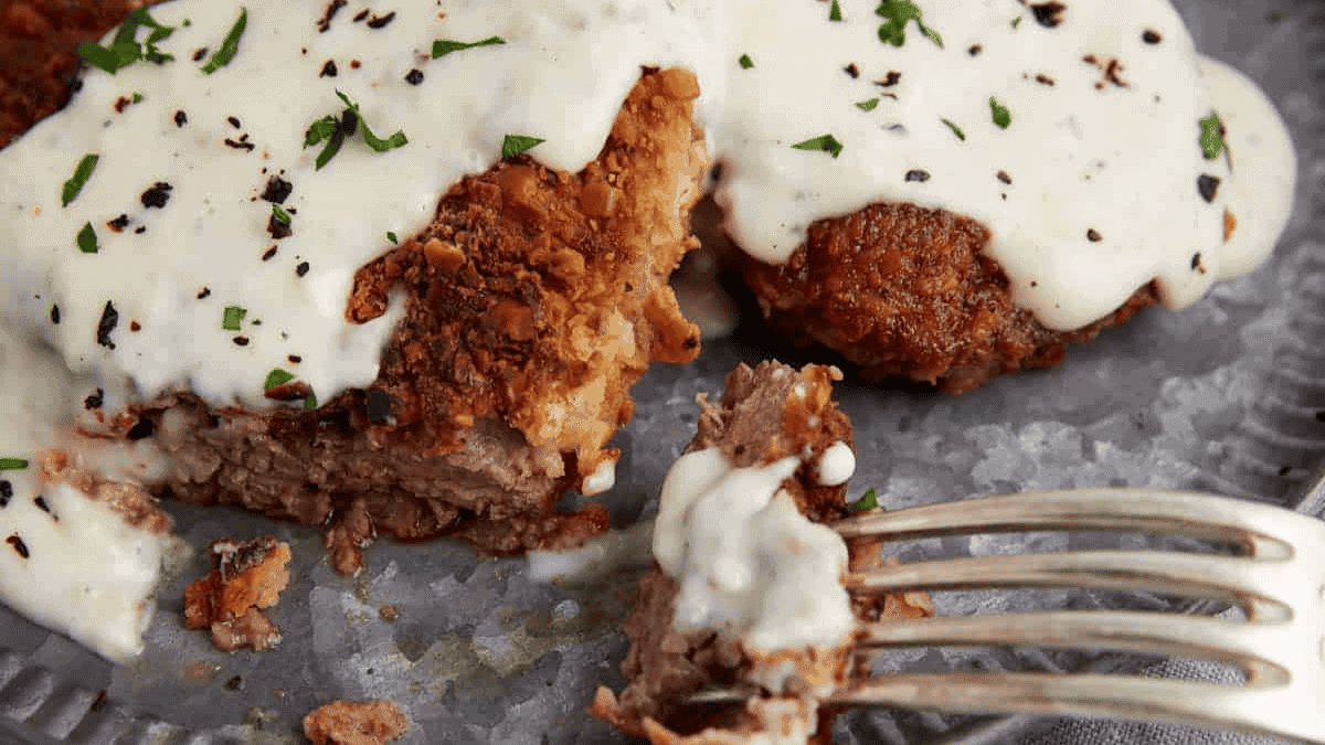 A close-up shot of crispy chicken fried steak topped with creamy gravy, garnished with chopped parsley and black pepper. One piece is partially cut, showing the inside, with a fork holding a small bite. The meal is served on a textured metal plate.