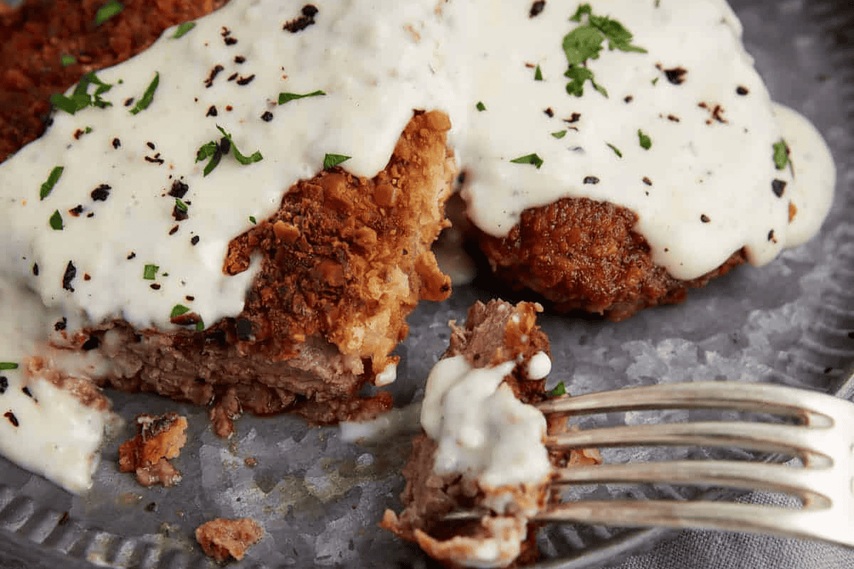 A close-up shot of crispy chicken fried steak topped with creamy gravy, garnished with chopped parsley and black pepper. One piece is partially cut, showing the inside, with a fork holding a small bite. The meal is served on a textured metal plate.