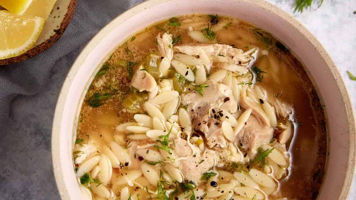 A bowl of orzo chicken soup with lemon slices next to it.