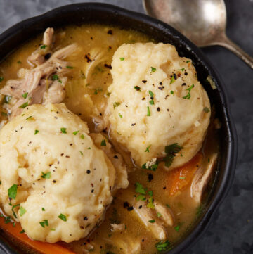 A bowl of chicken and dumplings. The dish features shredded chicken, carrots, and two large dumplings in a thick broth, garnished with chopped parsley and freshly ground black pepper. A silver spoon rests beside the bowl on a dark, textured surface.