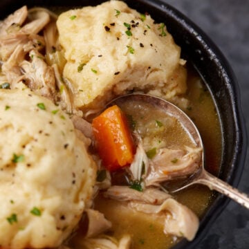 A close-up of a bowl of chicken and dumplings, featuring tender shredded chicken, chunks of carrots, and fluffy dumplings in a savory broth. The dish is garnished with freshly ground black pepper and chopped herbs. A spoon rests in the bowl, ready to serve.