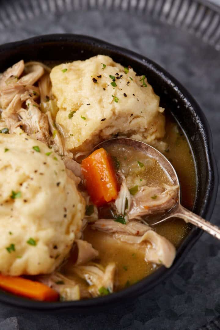 A close-up of a bowl of chicken and dumplings, featuring tender shredded chicken, chunks of carrots, and fluffy dumplings in a savory broth. The dish is garnished with freshly ground black pepper and chopped herbs. A spoon rests in the bowl, ready to serve.