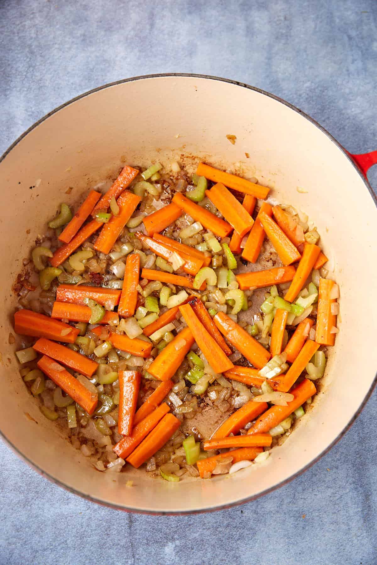 A large pot with red handles sits on a gray surface, filled with sautéed vegetables. The mix includes orange carrot sticks, chopped celery, and diced onions, all cooking and slightly caramelized. The vegetables are spread evenly across the bottom of the pot.