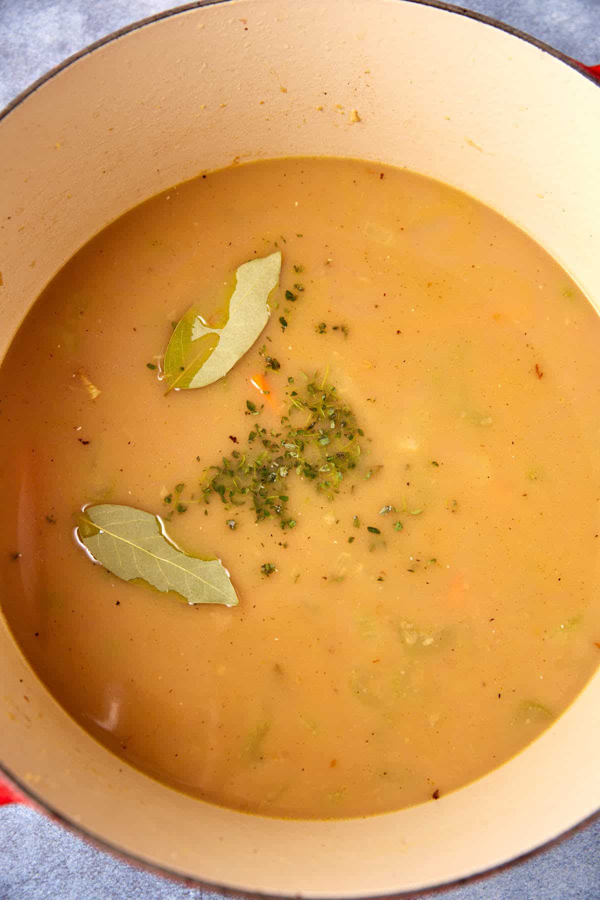 A large pot of creamy beige soup with visible chunks of vegetables. The soup is garnished with two bay leaves and a small sprinkling of finely chopped herbs in the center. The interior of the pot is off-white, contrasting with the soup's color.