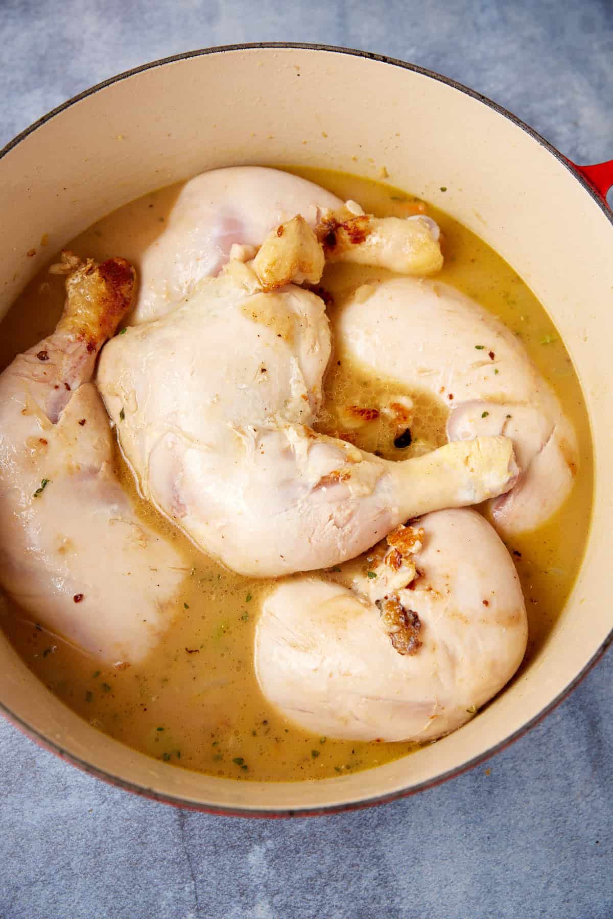 A red pot holds several pieces of partially cooked chicken in a light, creamy broth. The chicken pieces appear slightly browned around the edges, and the broth features visible herbs and seasonings. The container is situated on a blueish-gray surface.