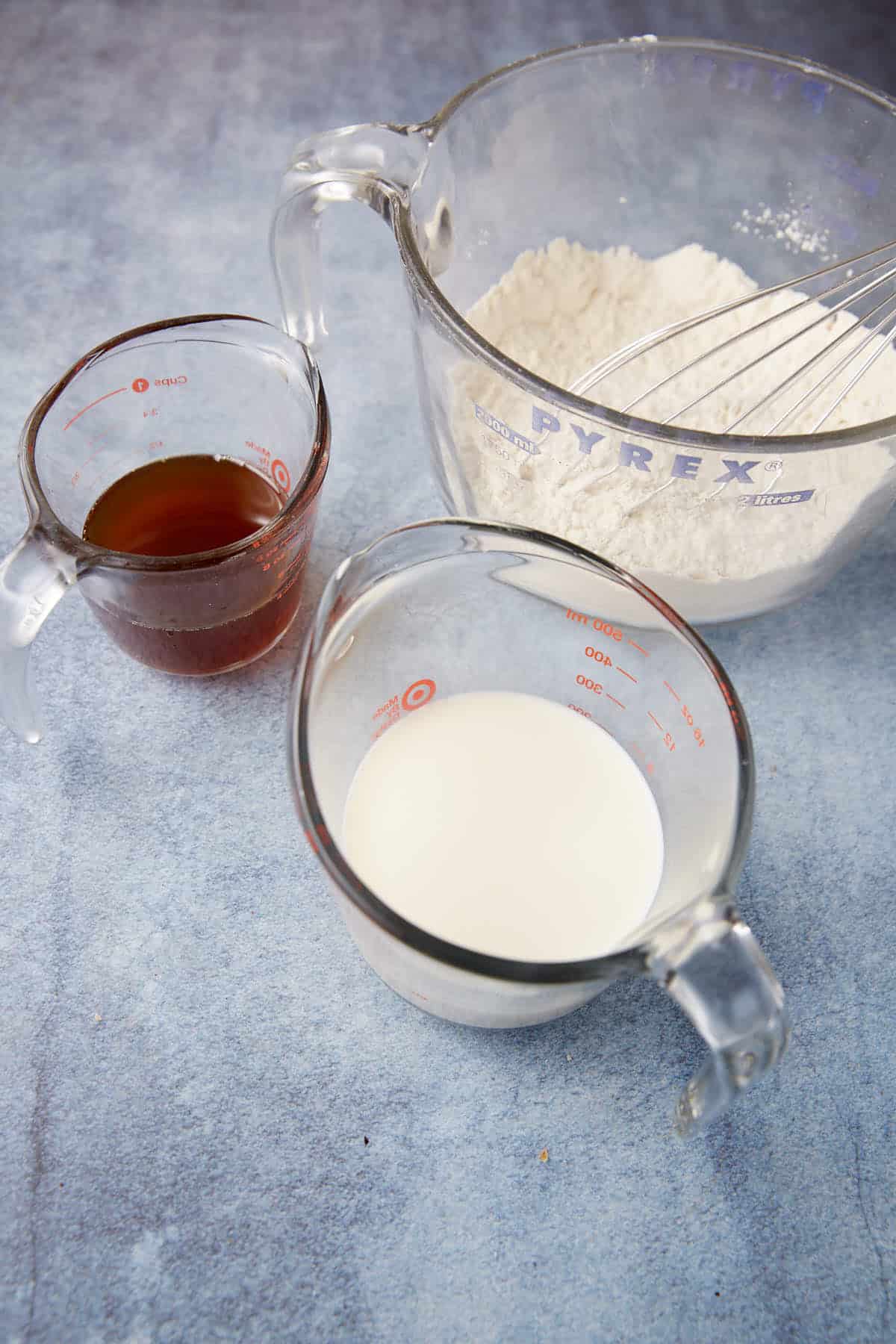 Three measuring cups are placed on a blue-gray surface. The large cup holds flour with a whisk inside it, the medium cup contains a brown liquid, and the small cup has a white liquid, likely milk. All measuring cups are made of clear Pyrex glass.