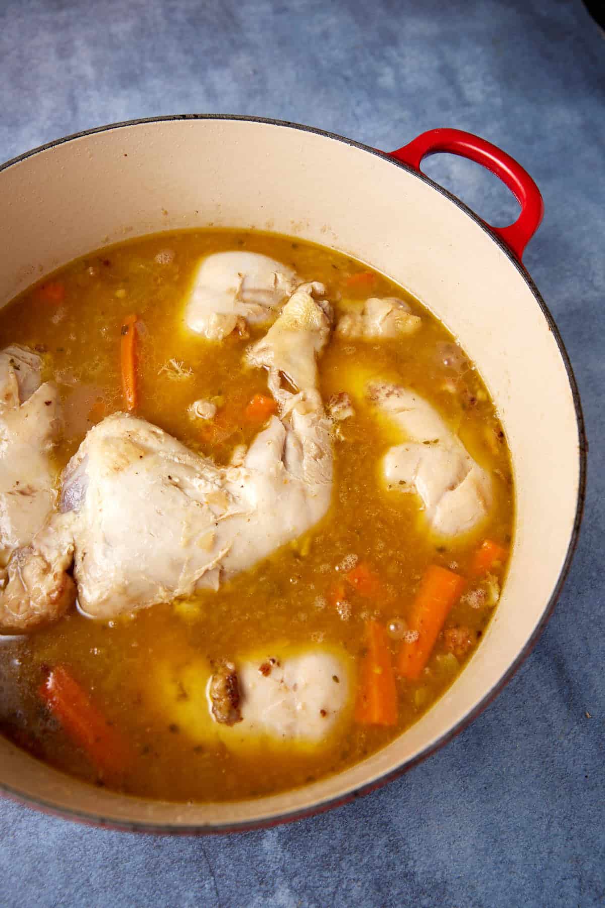 A red-handled pot filled with chicken pieces, carrots, and a golden broth, resting on a blue-grey surface. The contents are stewing, with visible herbs and spices in the liquid, indicating a flavorful cooking process.