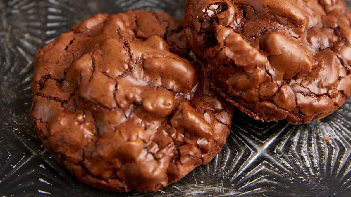 Close-up of two chocolate cookies with a cracked surface resting on a textured, metallic tray. The cookies appear chewy with chunks of chocolate embedded in them. In the background, there are additional cookies and a glass of milk atop a white napkin.