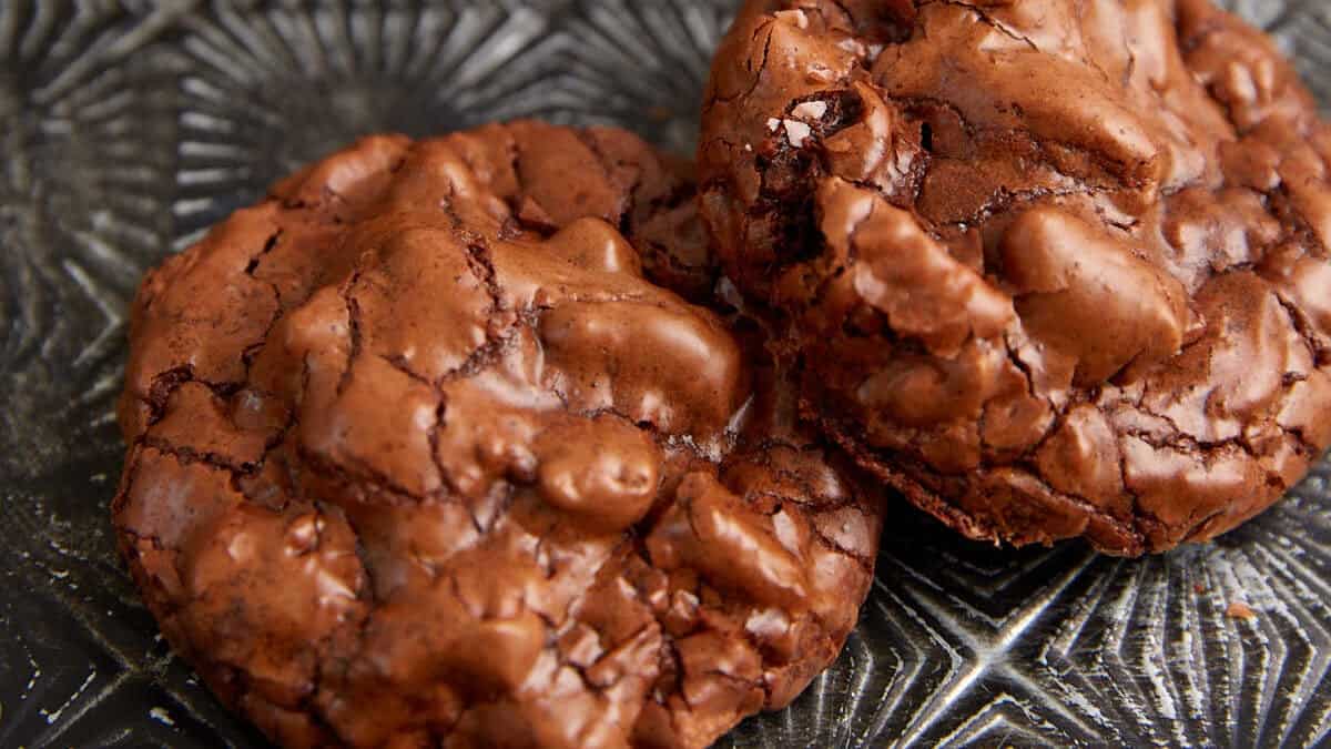 Close-up of two chocolate cookies with a cracked, glossy surface on a textured, dark-colored plate. The cookies have a rich, fudgy appearance with visible pieces of chocolate embedded within them.