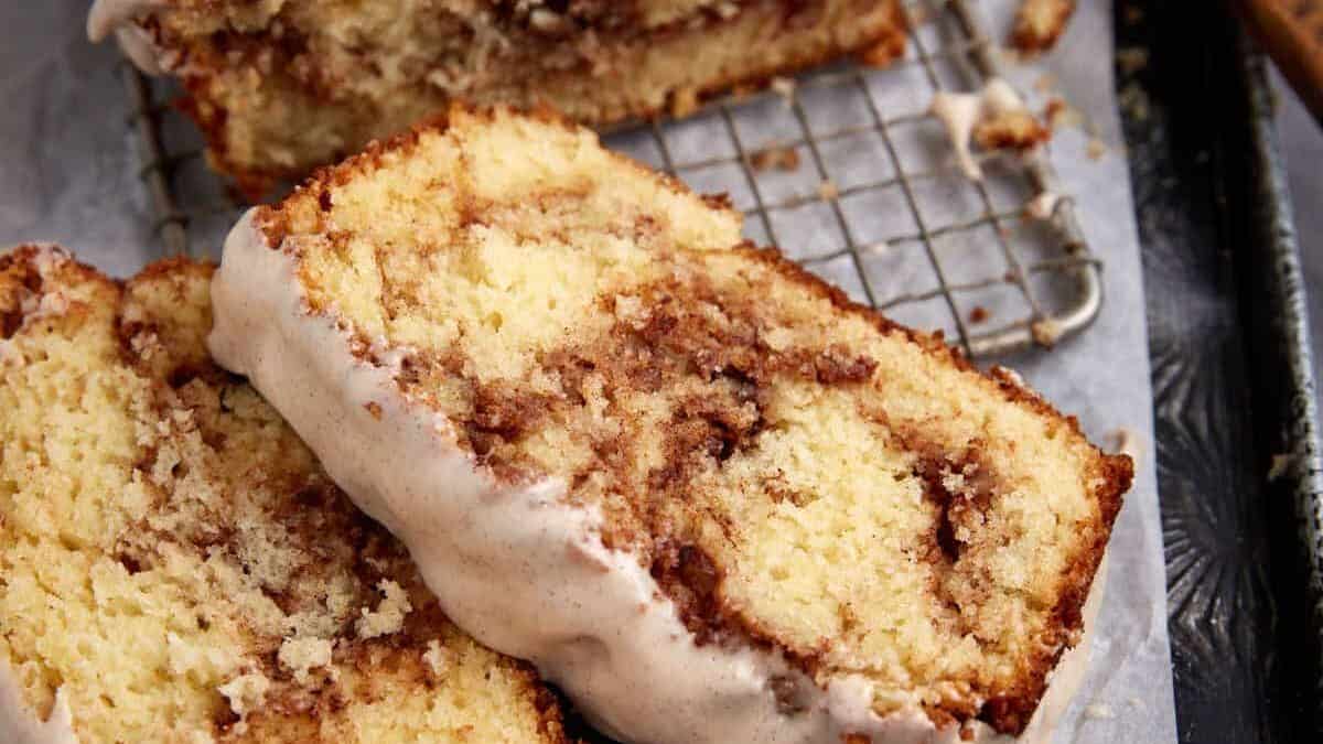 Close-up of two slices of cinnamon swirl bread on a cooling rack. The bread is light golden in color with visible cinnamon swirls and topped with a layer of white glaze. A loaf sits partially in the background. Dark and rustic surface underneath.
