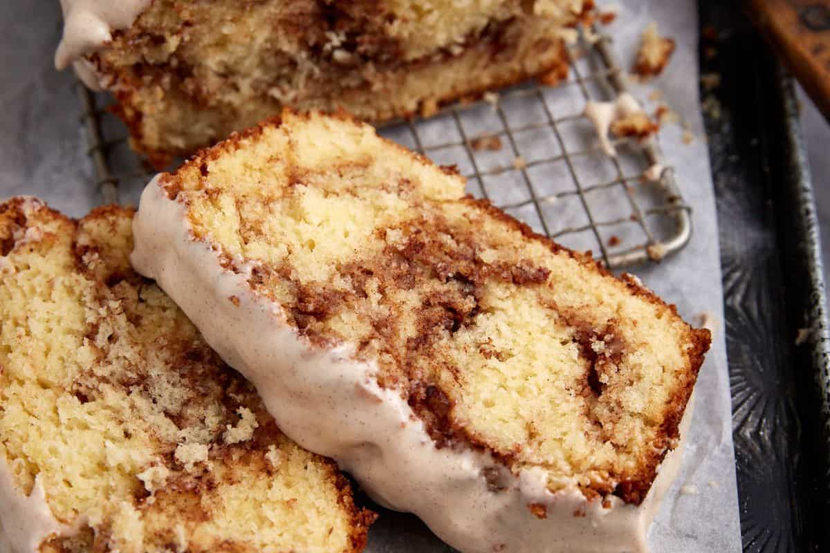 Close-up of two slices of cinnamon swirl bread on a cooling rack. The bread is light golden in color with visible cinnamon swirls and topped with a layer of white glaze. A loaf sits partially in the background. Dark and rustic surface underneath.