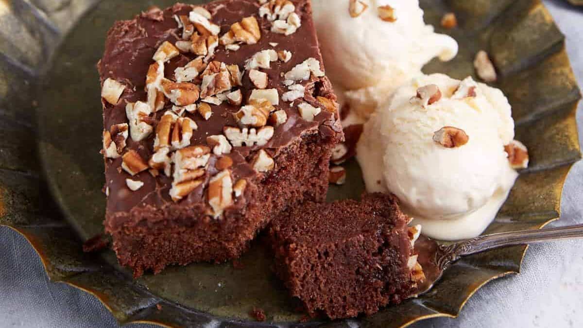 A metal plate holds a piece of chocolate sheet cake topped with chopped nuts, served alongside two scoops of vanilla ice cream also sprinkled with nuts—a perfect back to school dessert. In the background, there's a baking tray with more chocolate cake and two bottles of Coca-Cola.
