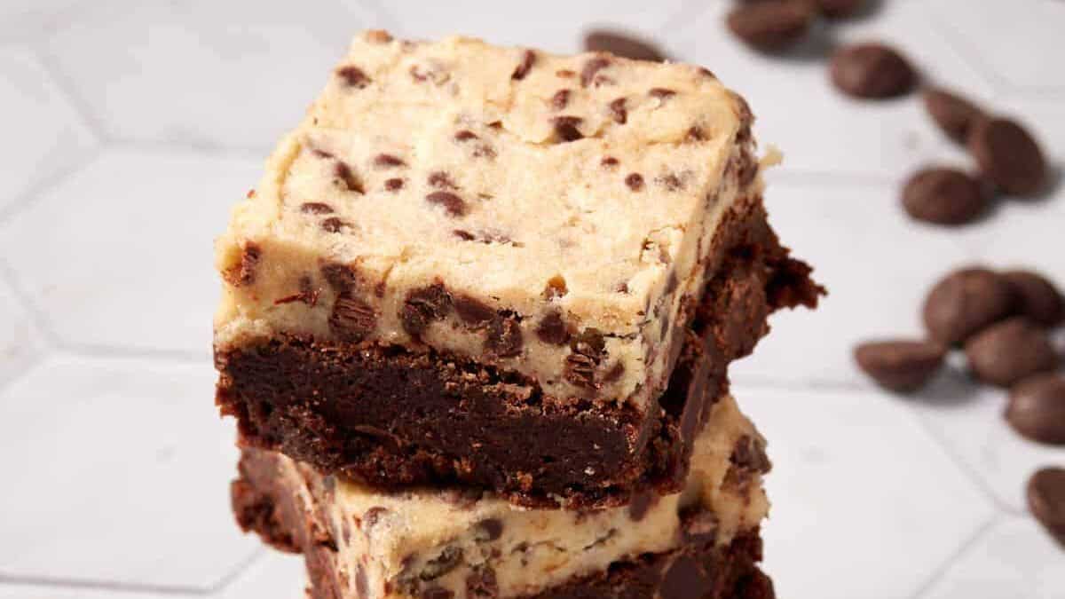 Two stacked dessert bars with a dense chocolate brownie base and a thick layer of chocolate chip cookie dough on top, perfect for back to school desserts, with scattered chocolate chips in the background.