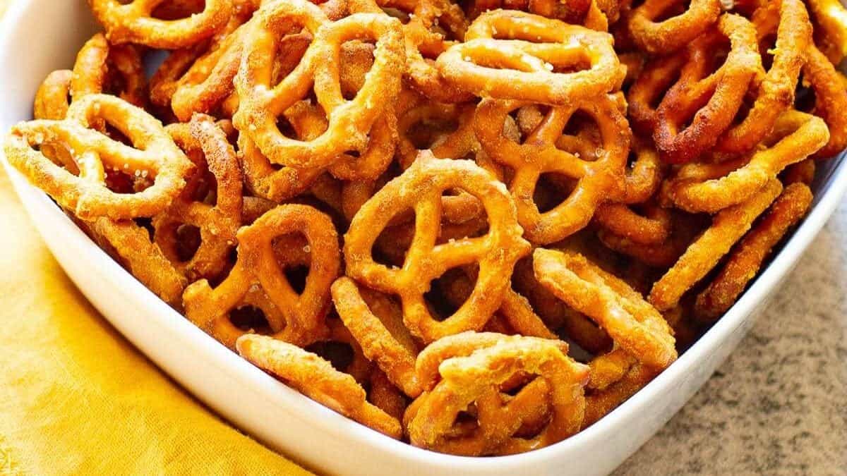 A white square bowl filled with seasoned pretzel pieces is placed on a yellow cloth. The pretzels are golden brown with visible seasoning, suggesting a savory flavor. The background is a light-colored surface, enhancing the vibrant colors of the cloth and pretzels.