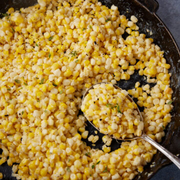A skillet filled with creamy garlic parmesan corn, featuring kernels of corn mixed with a creamy sauce and garnished with black pepper and chopped herbs. A spoon is scooping some corn from the skillet.