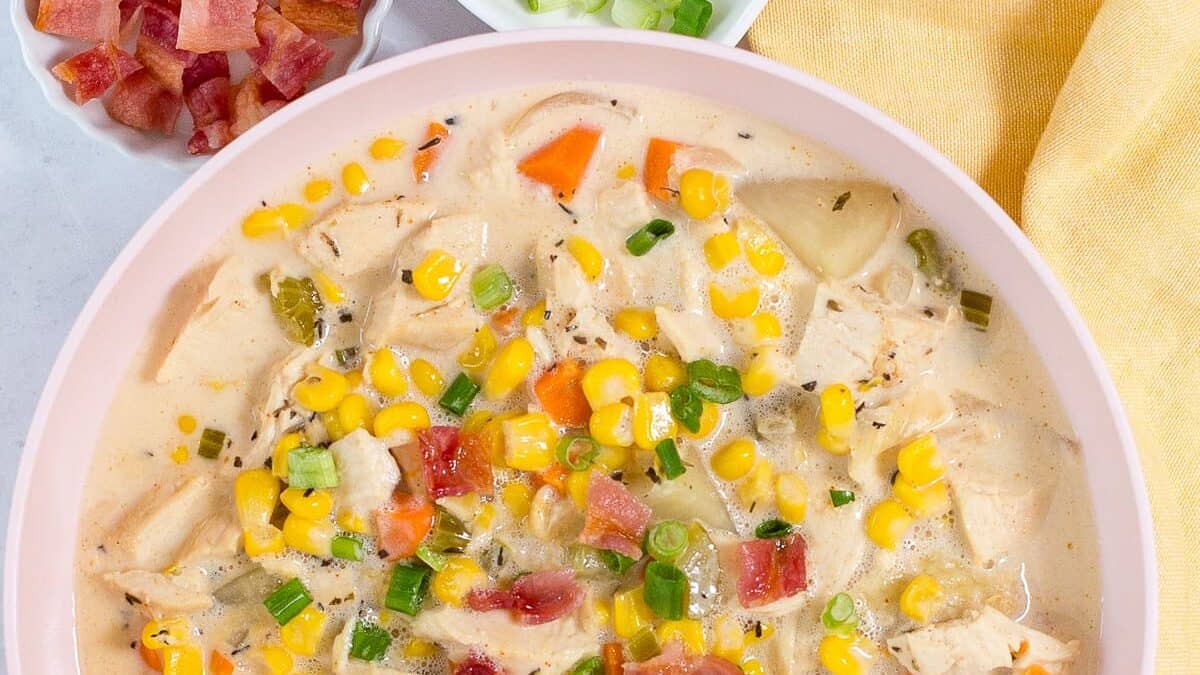A bowl of creamy chicken corn chowder garnished with chopped bacon and green onions. The soup is filled with chunks of chicken, corn kernels, diced carrots, and celery in a creamy broth. A small bowl of bacon bits and a dish of sliced green onions are in the background.