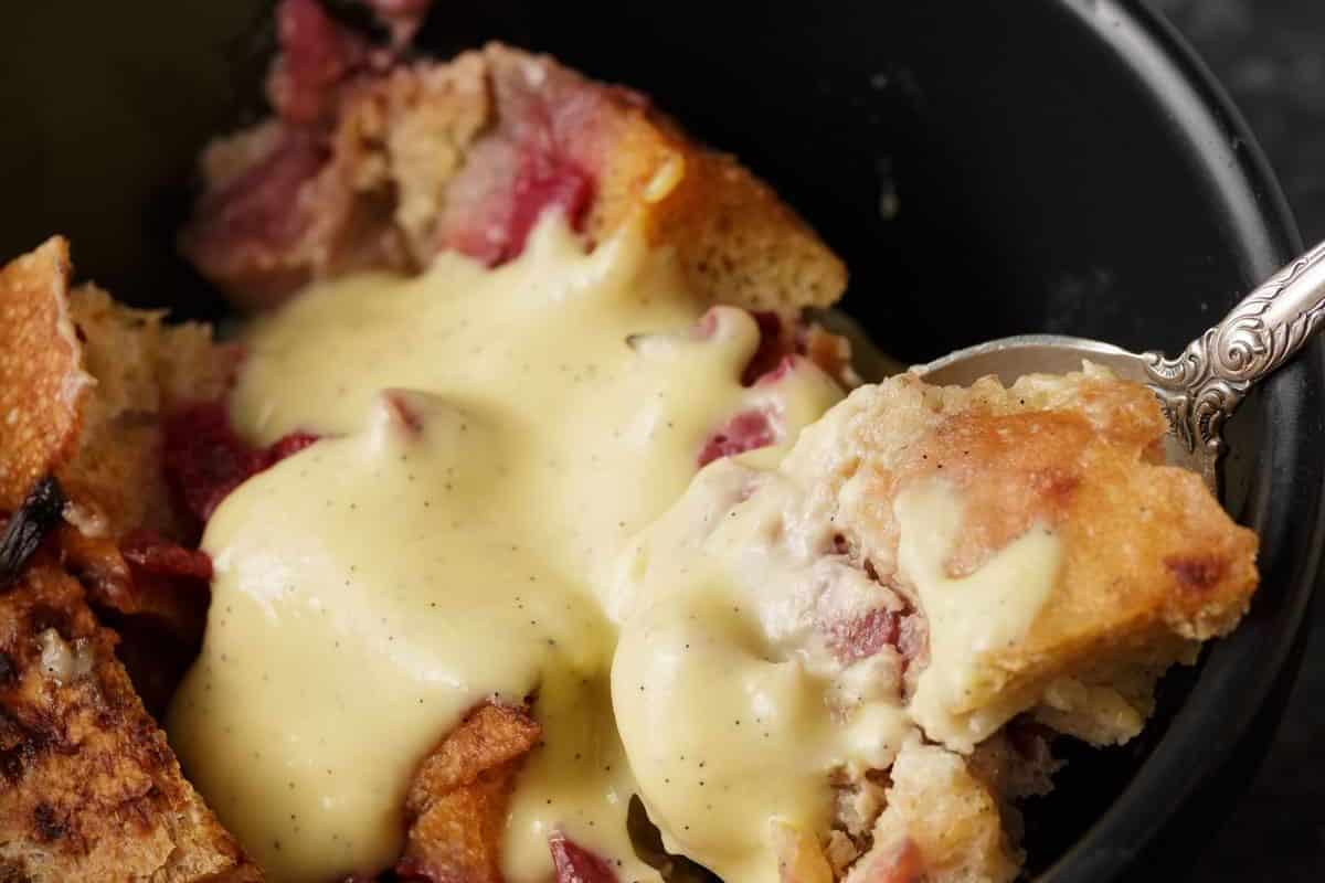 A close-up view of a bread pudding topped with a generous amount of creamy vanilla sauce. The pudding consists of bread pieces and visible chunks of red fruit, likely berries, served in a black bowl with a spoon scooping a portion.