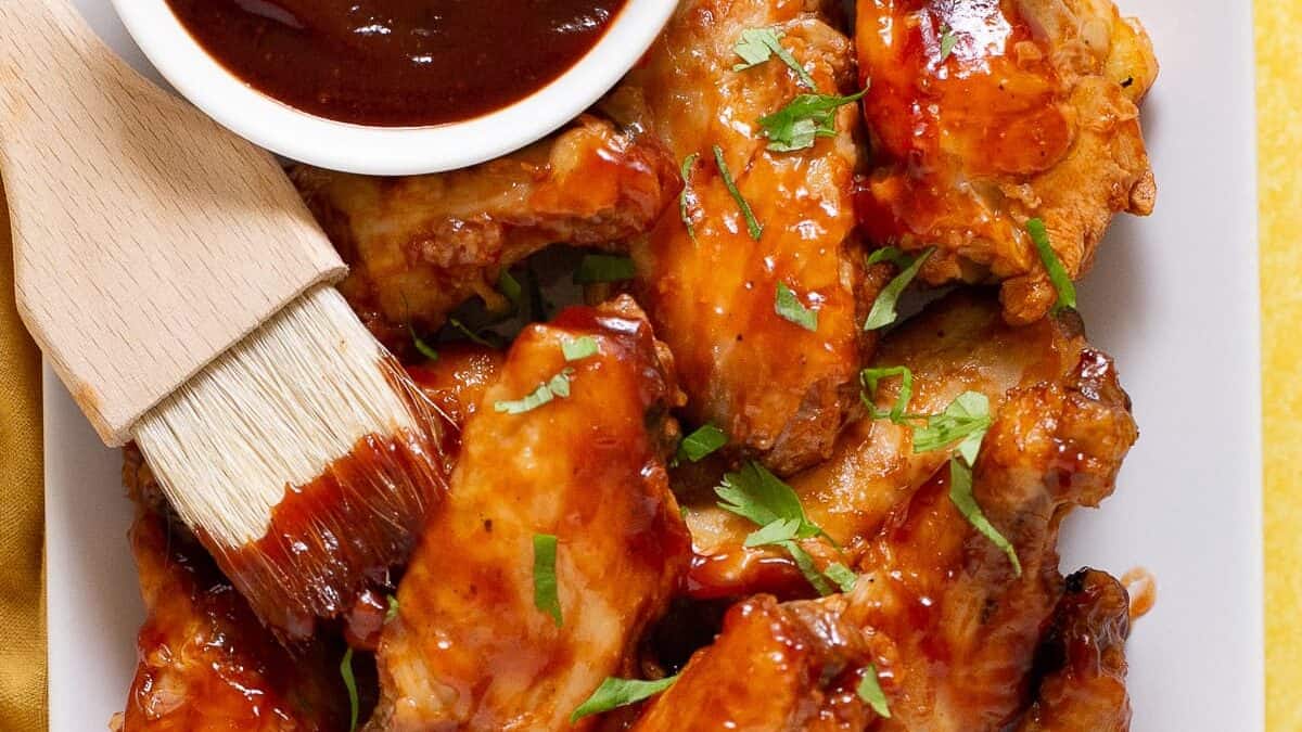 A rectangular white plate filled with saucy barbecue chicken wings garnished with chopped herbs. A small dish of barbecue sauce and a sauce brush are placed at the corner of the plate. A yellow napkin is partially visible on the left side.