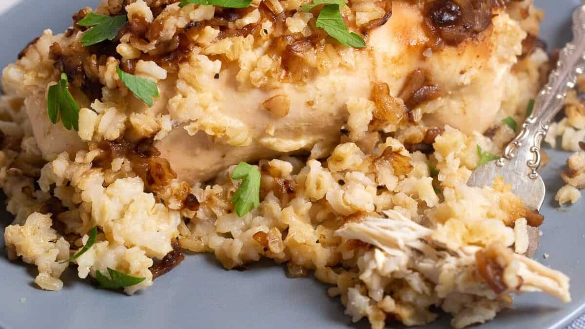 A close-up of a plate featuring a serving of baked chicken covered in caramelized onions and garnished with chopped parsley. The chicken is surrounded by seasoned rice. In the background, there are blurred onions and a striped cloth.
