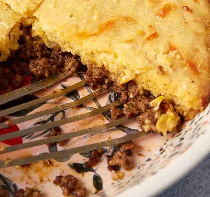 A round ceramic dish with a floral pattern holds a partially eaten hamburger pie, featuring a mashed potato topping over a savory meat filling. A metal spatula rests in the dish. The background includes a navy blue cloth and a wooden board.