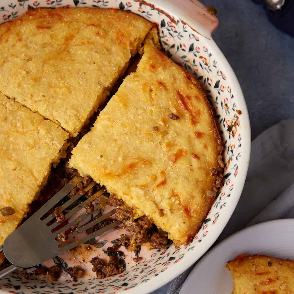 A round dish filled with a baked savory hamburger pie featuring a golden-brown crust and a visible meat filling. A triangular slice has been cut and a spatula is lifting it out. The dish has a floral pattern on its rim, and part of the pie is served on a plate nearby.