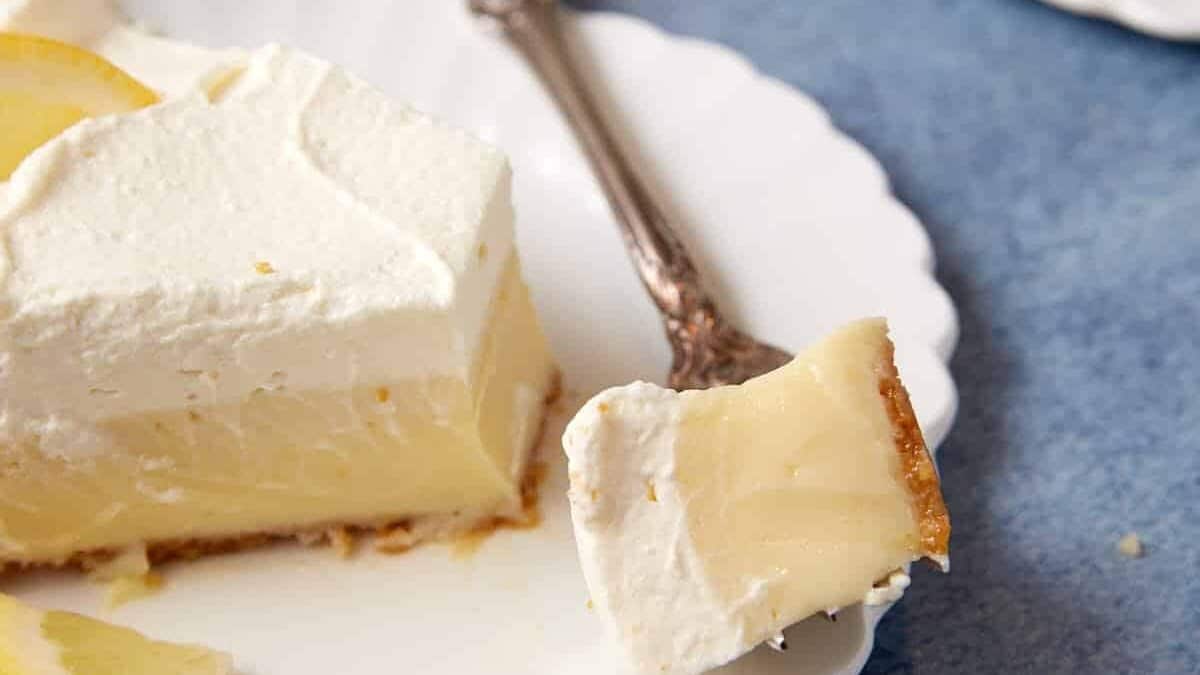 A slice of creamy lemon cheesecake sits on a white plate with a decorative fork. A bite-sized piece of the cheesecake is on the fork, accompanied by small lemon slices on the plate, reminiscent of a classic lemon icebox pie. The background features a blue cloth and another plate with a cheesecake slice.