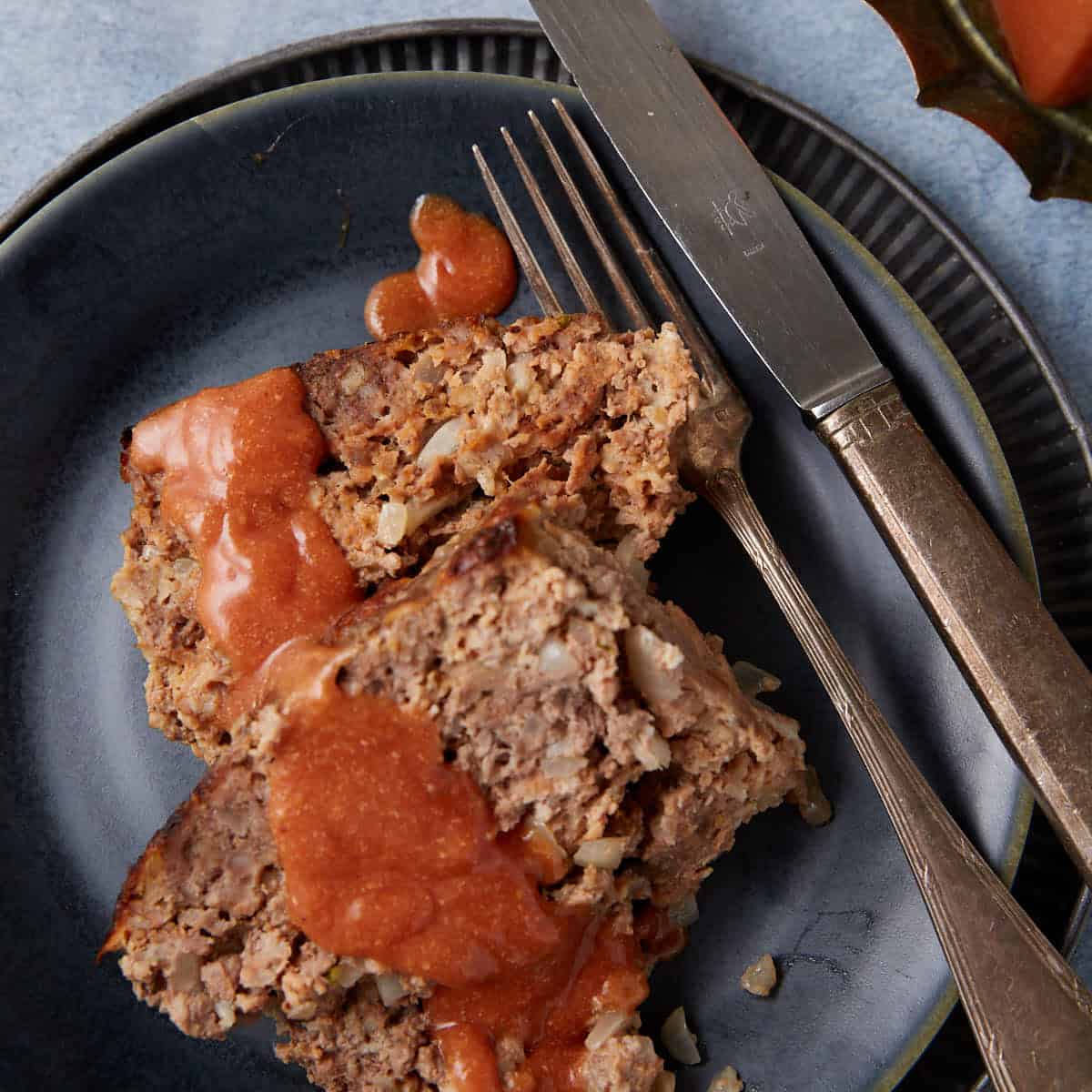 A close-up of a plate with slices of meatloaf, topped with a tomato-based sauce. The meatloaf is garnished with visible onion bits and is served with a fork and knife placed beside it. The plate is set on a textured surface and is part of a rustic table setting.