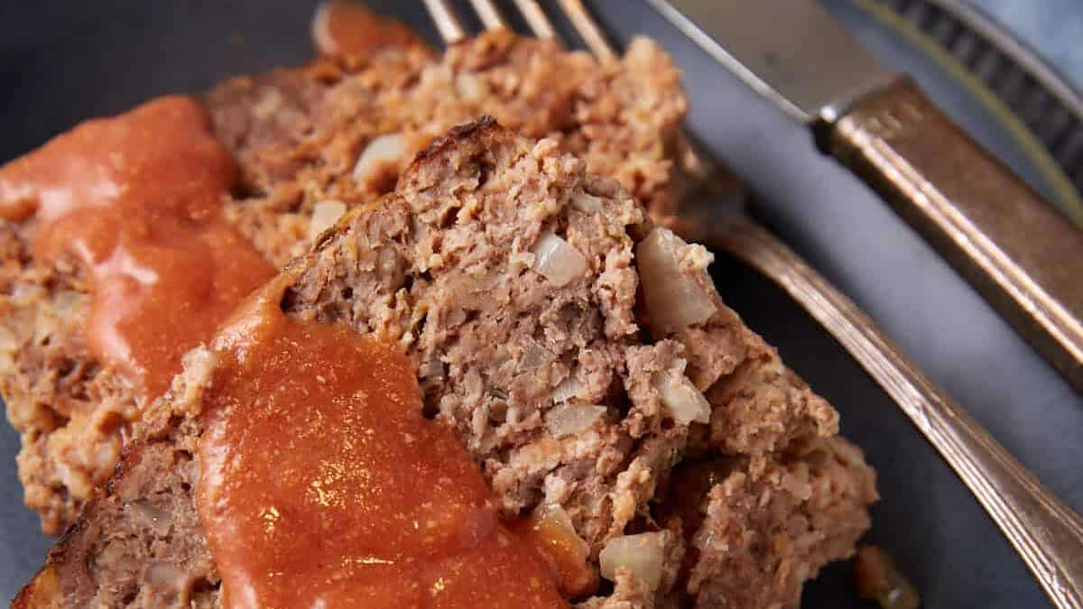 A close-up of a blue plate with succulent meatloaf slices drizzled in red sauce. A fork and knife are artfully placed beside the meatloaf on the plate, all set against a light blue table surface.
