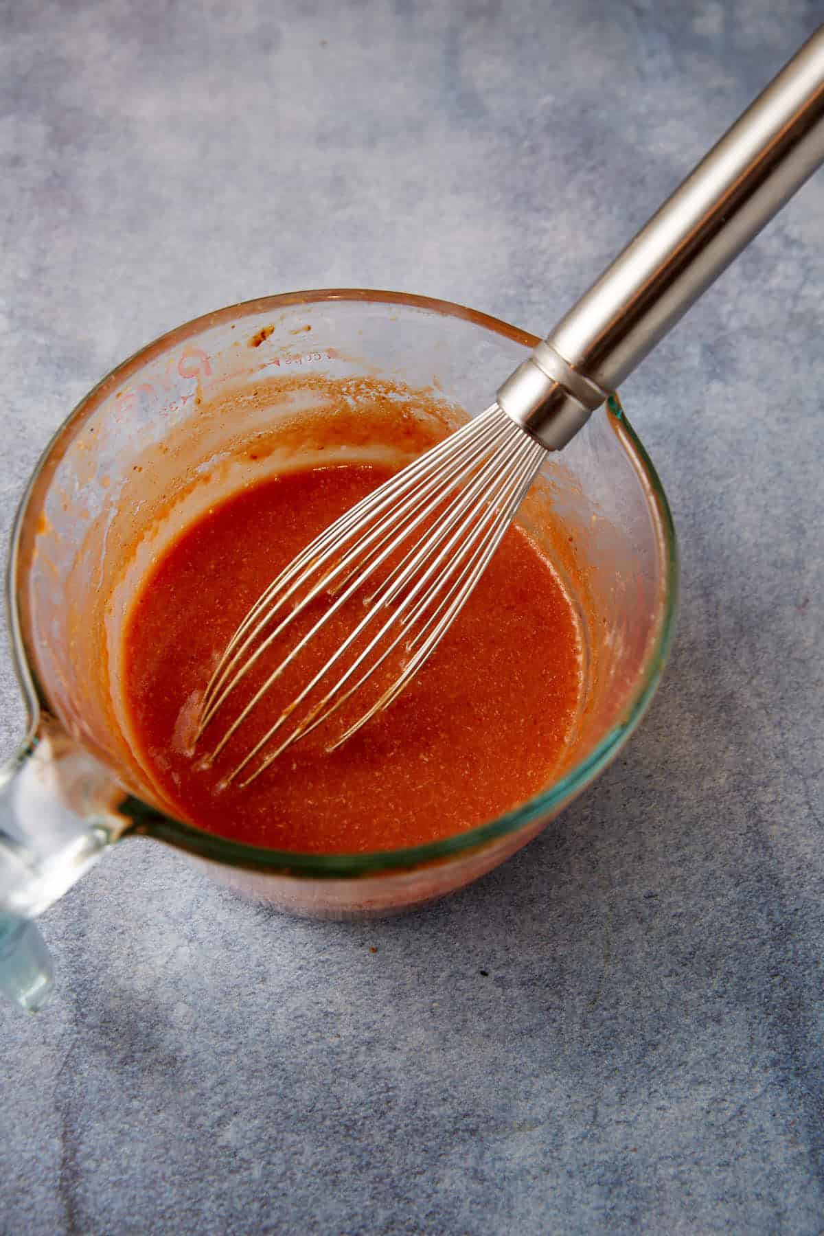 A glass measuring cup filled with a reddish sauce being whisked with a metal whisk. The cup is placed on a gray surface. The mixture appears to be well-mixed, making it perfect for enhancing your meatloaf recipe or other cooking and baking creations.