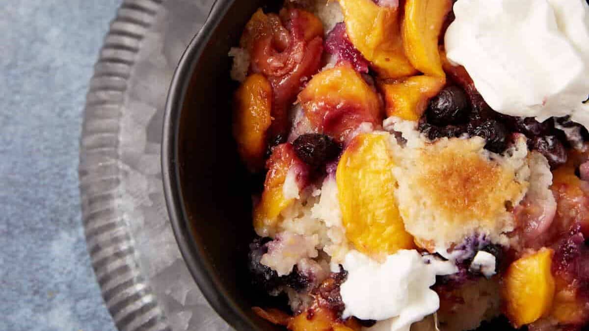 A bowl of peach and berry cobbler topped with whipped cream is on a dark plate with a spoon. The cobbler consists of baked peaches, berries, and a golden crust. In the background, part of a baking dish filled with more cobbler and a whole peach are visible.