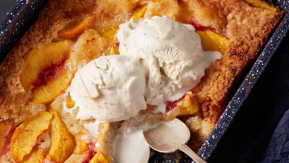 A blue baking pan with peach cobbler cake covered with ice cream.