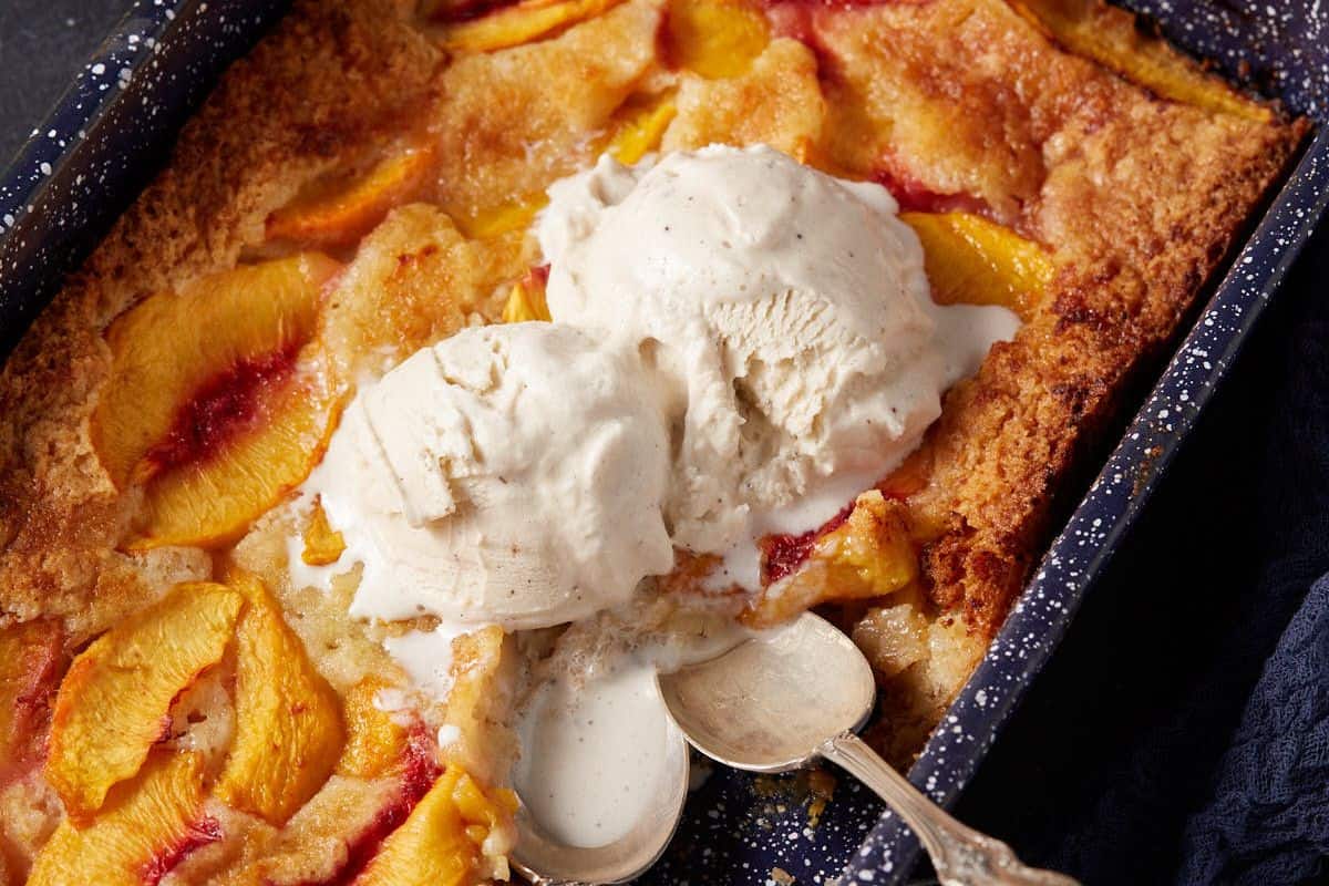 A close-up of a peach cobbler served in a dark baking dish. The cobbler, a southern dessert classic, has golden-brown crusts and visible peach slices. Two scoops of melting vanilla ice cream are placed on top, with a silver spoon resting beside them.