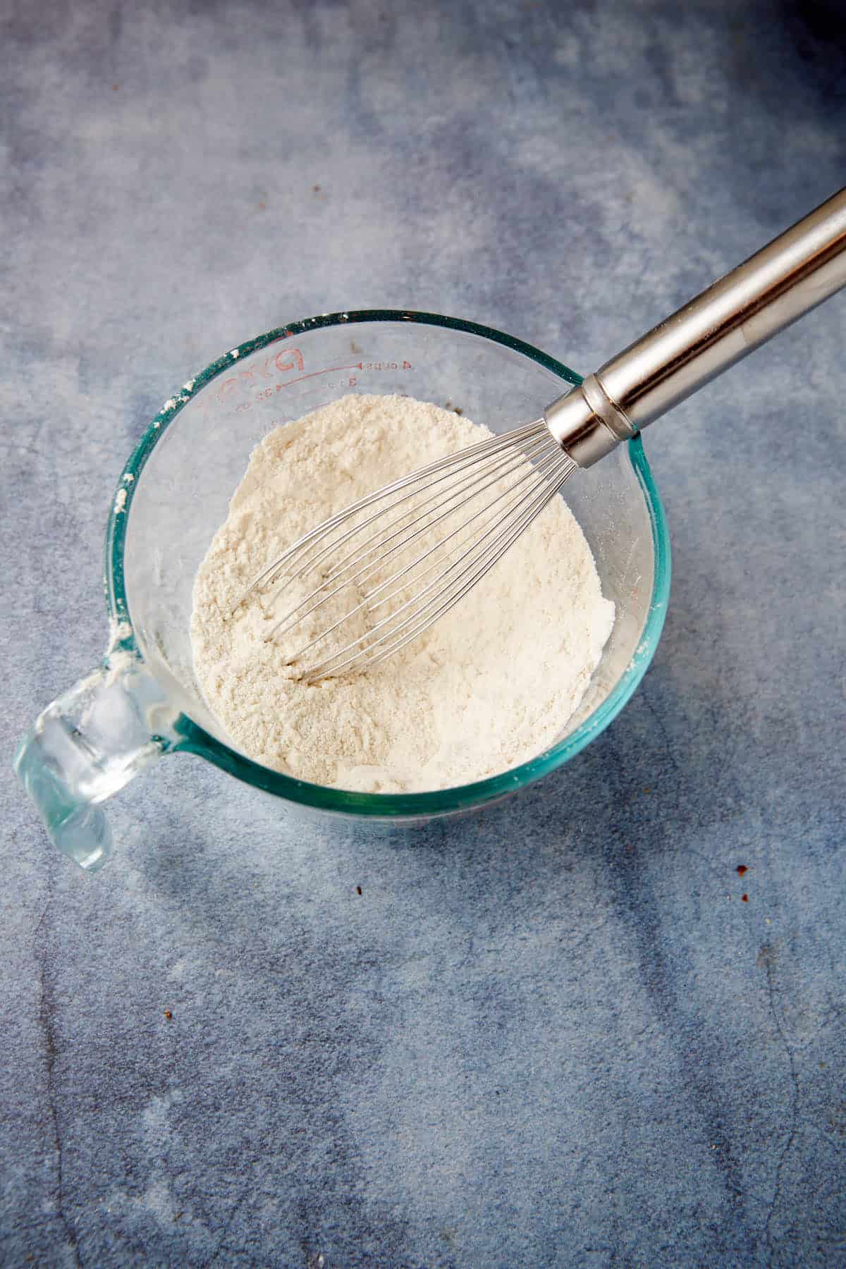 A glass measuring cup filled with dry ingredients and a metal whisk resting inside it, ready to create the perfect peach cornbread muffins, sits on a textured, light gray surface.