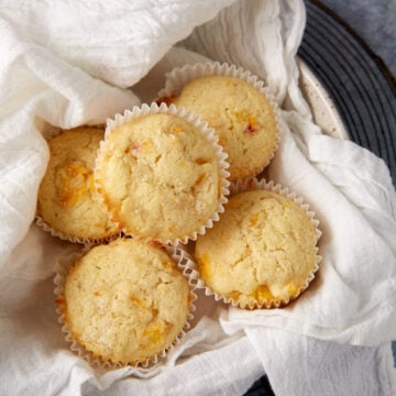 A close-up of five homemade peach cornbread muffins nestled in a white cloth-lined basket. The muffins have a golden-brown exterior with visible bits of peach, suggesting they are fruit-flavored, and are placed in white paper liners. The basket is on a dark surface.