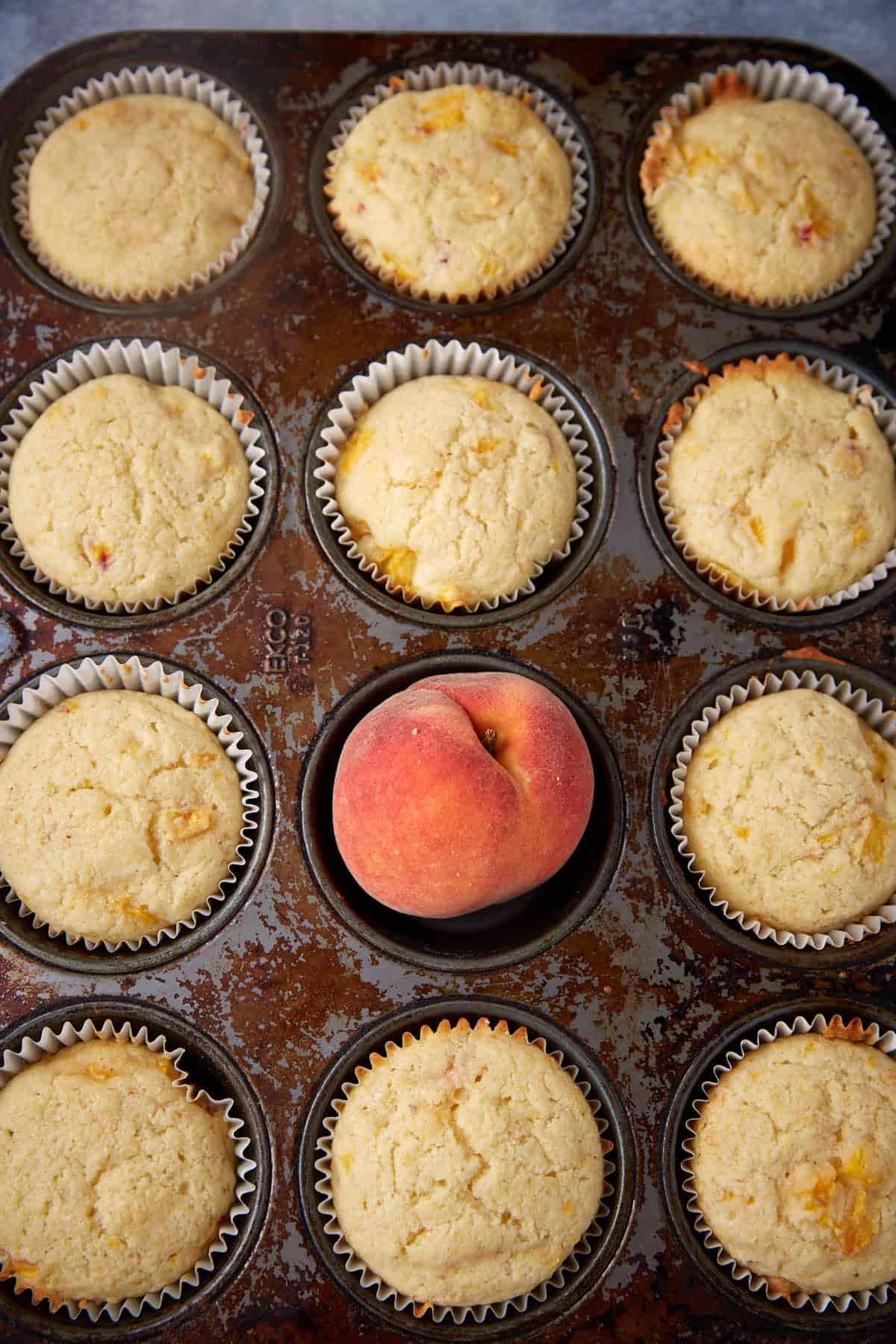 A muffin tray with 12 slots is filled with 11 baked, golden-brown peach cornbread muffins in paper liners. One slot contains a whole peach instead of a muffin. The tray has a rustic, slightly worn appearance.
