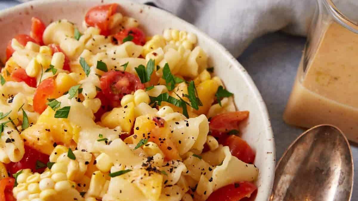 A bowl of peach pasta salad with dressing in a pticher on the side.