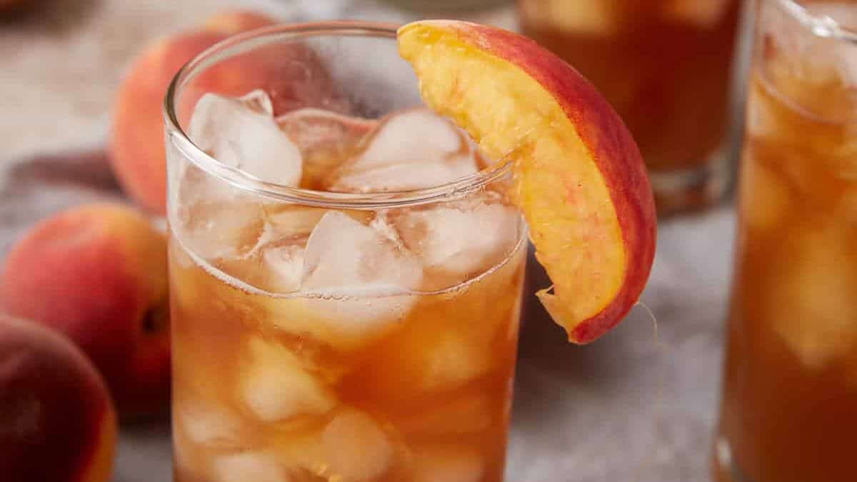 A close-up of a refreshing iced tea in a glass, garnished with a peach slice. The beverage is filled with ice cubes, and there are additional glasses and a pitcher in the background, along with some whole peaches placed casually around the setup.