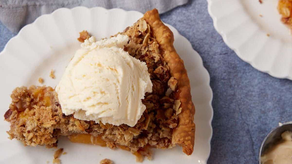 A slice of fruit pie with a crumbly topping is served on a white scalloped plate. The pie is topped with a scoop of vanilla ice cream. A portion of another pie slice is visible on another plate in the background, with a light gray cloth nearby.