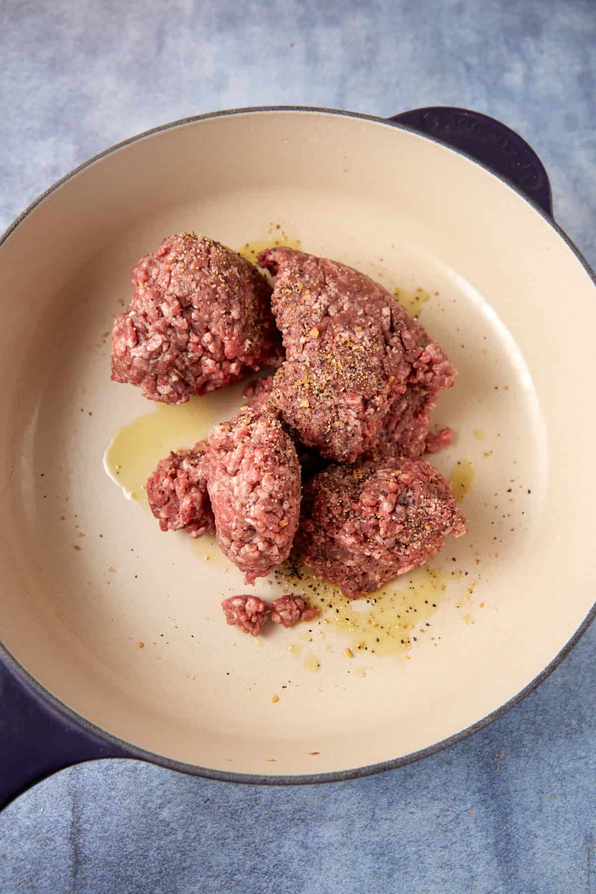A round, cream-colored pan with dark handles contains several portions of raw ground meat, destined for a delicious cheesesteak casserole, seasoned with pepper and drizzled with oil, set on a blue-toned surface.