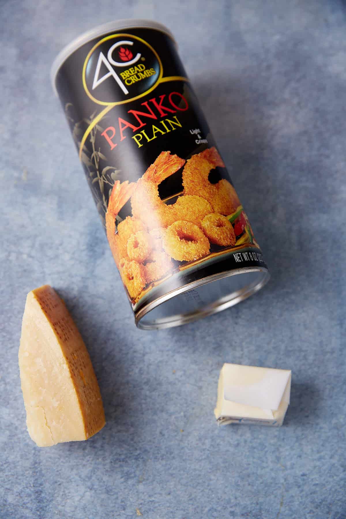 A canister of plain panko bread crumbs lies on its side on a blue-gray surface. In front of the canister, there is a wedge of Parmesan cheese and a small pat of butter, hinting at the perfect ingredients for a cheesesteak casserole. The canister features images of breaded fried food.