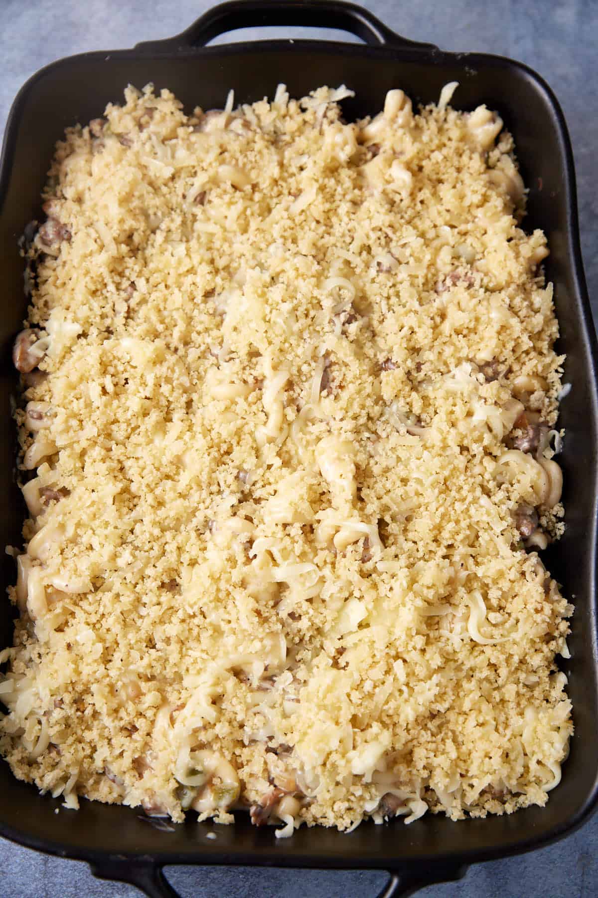 A rectangular baking dish filled with a creamy cheesesteak casserole topped with a golden, breadcrumb crust. The pasta is mixed with pieces of mushrooms, visible beneath the crispy topping. The dish contrasts beautifully against a neutral grey background.