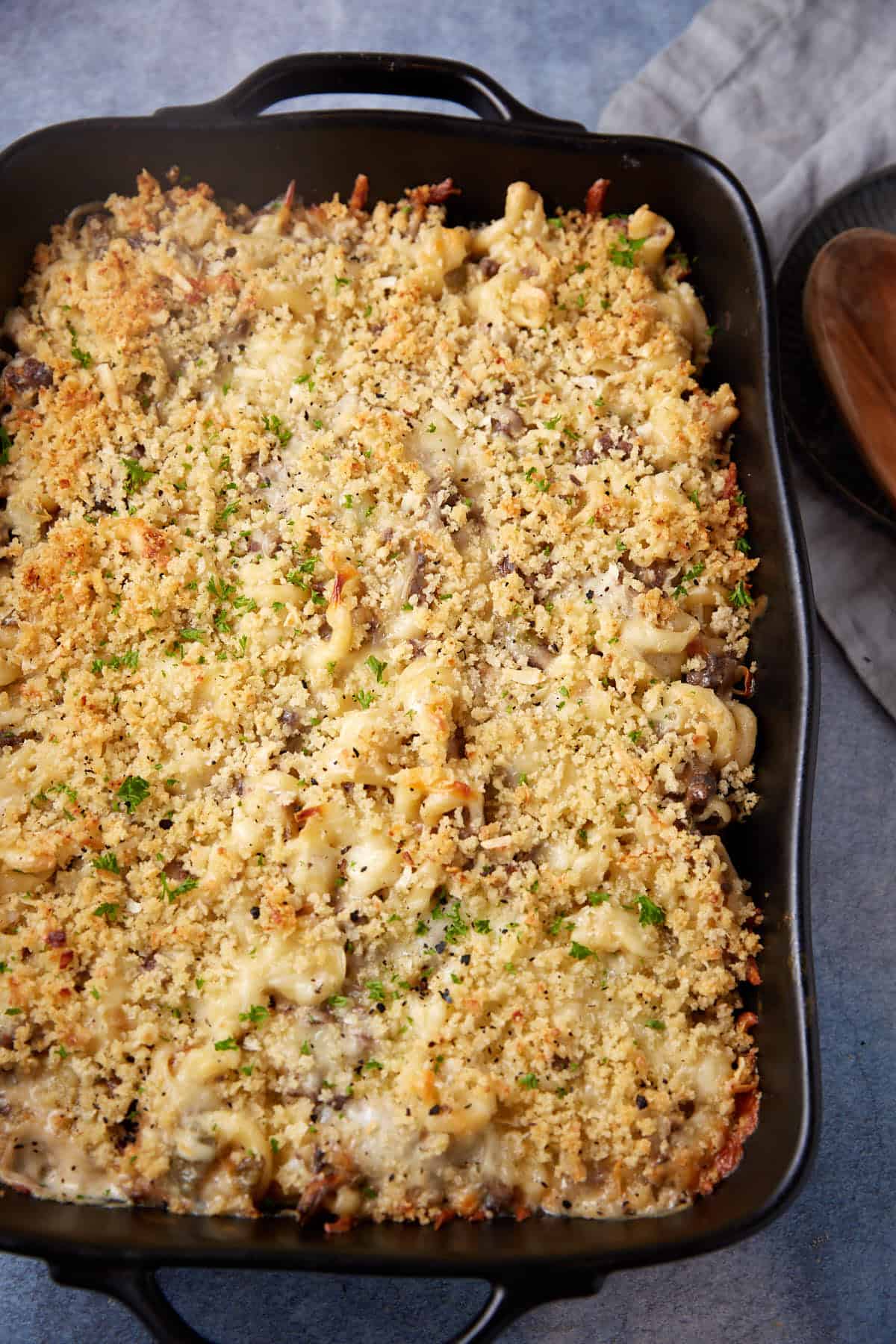 A rectangular baking dish filled with a golden brown, crispy-topped cheesesteak casserole sits on a grey surface. The dish appears to have a crumb topping with hints of herbs and melted cheese. A gray napkin is placed beside the casserole with a wooden spoon on a black plate.