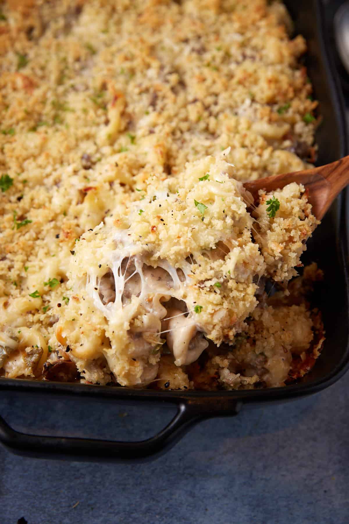 A dish of baked creamy cheesesteak casserole with a golden breadcrumb topping is shown. A serving spoon lifts a portion, revealing gooey melted cheese and tender pieces of chicken and rice. The casserole is garnished with chopped parsley, adding a touch of green.