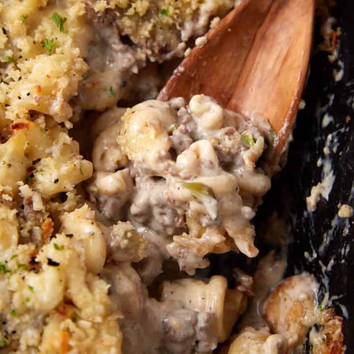 Close-up of a creamy beef and macaroni casserole with seasoned bread crumb topping. The mixture is being scooped with a wooden spoon, revealing tender pasta, ground beef, and a rich, cheesy sauce, accented with herbs and a crispy, golden-brown crust.