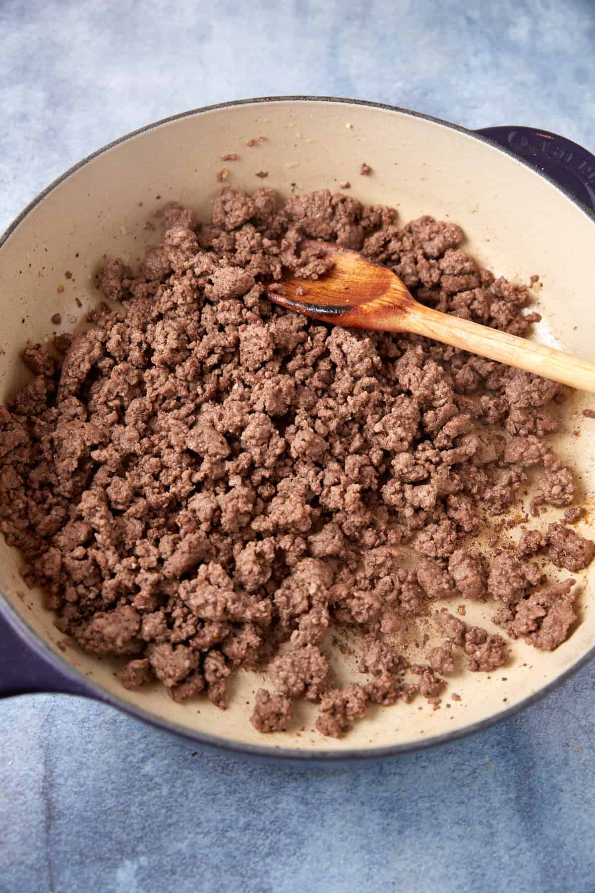 A skillet filled with cooked ground beef, being stirred with a wooden spoon on a gray countertop. The beef is browned and crumbled, indicating that it has been cooked thoroughly—just the perfect base for a savory cheesesteak casserole.